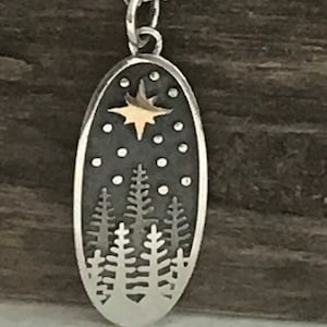 以下が含まれることがあります： 銀色のペンダントネックレス。銀色の楕円形のチャームには、金色の星と雪が降る森のシルエットが描かれています。