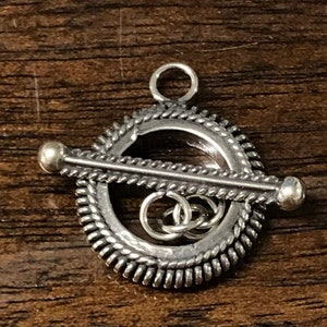 Op de afbeelding: Zilveren toggle-sluiting met een rond, gestructureerd ontwerp en een decoratieve stang met twee ronde kralen aan de uiteinden.