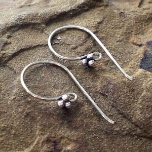 May include: A pair of silver earrings with a minimalist design. Each earring has a curved hook and a cluster of three small, round beads. The earrings are shown on a textured, brown surface.
