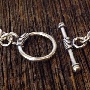 Op de afbeelding: Een zilveren kettingarmband met een toggle-sluiting. De sluiting is gemaakt van een gedraaide metalen staaf met een ronde ring.