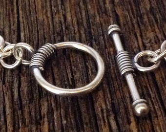 May include: A silver chain bracelet with a toggle clasp. The clasp is made of a twisted metal bar with a round ring.