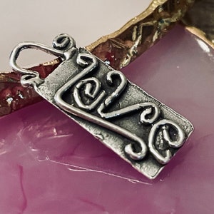 May include: A silver pendant with the word "love" in a cursive, swirly font. The pendant is rectangular with a textured surface and a small loop at the top for a chain. The background is a pink and gold surface.