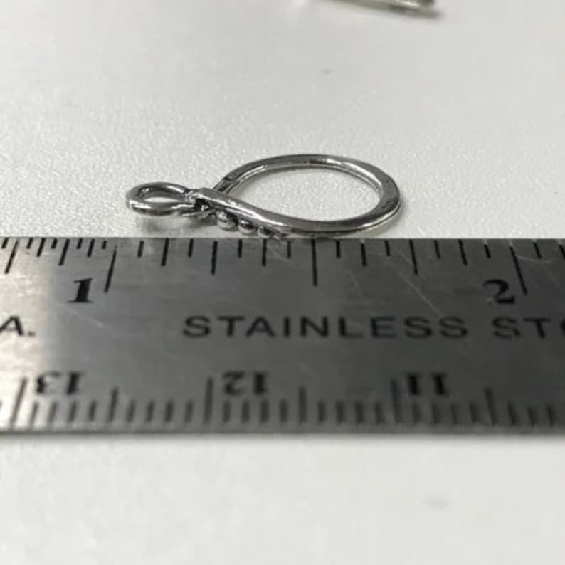 May include: A silver-toned metal jewelry finding with a small loop and a decorative bead detail. The finding is measured against a ruler, showing it is approximately 1 inch in length.