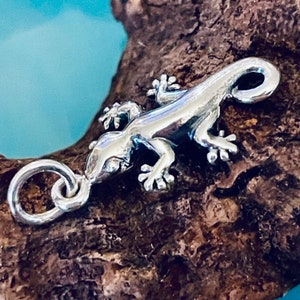 Op de afbeelding: Een zilveren bedel in de vorm van een gekko. De bedel heeft een lus om aan een armband of ketting te bevestigen.