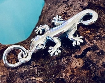 May include: A silver charm in the shape of a gecko lizard. The charm has a loop for attaching to a bracelet or necklace.