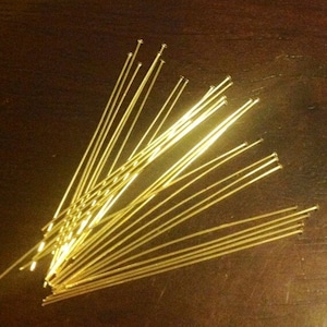 May include: A collection of gold-colored head pins, each with a small, rounded head and a long, slender wire shaft. The pins are scattered across a dark brown surface, with some overlapping. These pins are likely used for jewelry making or crafting.