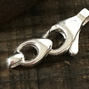 May include: Close-up of a shiny silver jewelry clasp. The clasp is composed of two interlocking parts, each with a rounded design. The metal has a polished finish, reflecting light. The background is a dark, textured surface.