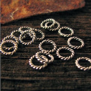 Puede incluir: Primer plano de varios pequeños anillos de salto plateados con un diseño de cuerda trenzada. Los anillos están dispersos sobre una superficie de madera oscura y texturizada, con un trozo de material marrón en el fondo. Los anillos son probablemente para la fabricación de joyas.