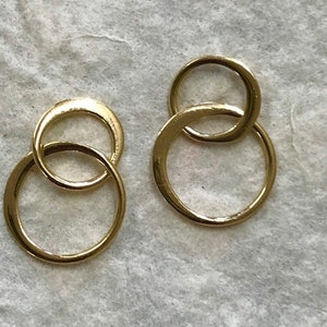 Puede incluir: Un par de pendientes de color dorado con un diseño de doble círculo. Los círculos están entrelazados, creando un aspecto moderno y minimalista.