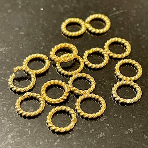 Puede incluir: Una colección de 12 anillos de salto de metal de color dorado con un diseño de cuerda trenzada. Los anillos son todos del mismo tamaño y están dispuestos en un patrón aleatorio.