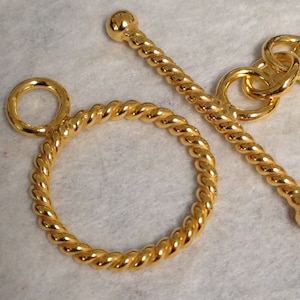 May include: Close-up of a gold-colored toggle clasp. The clasp features a circular ring and a bar with a ball end, both with a twisted rope design. The jewelry component is set against a light background.