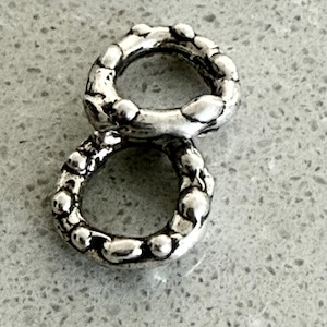May include: Two interconnected silver-toned rings with a textured, bumpy surface. The metal has a polished finish with dark recessed areas between the raised, rounded nodules that form the circular structure of each individual ring component.