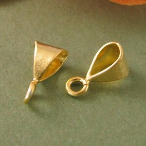 Puede incluir: Dos baúles de colgante de metal dorado con un acabado liso y cepillado. Los baúles tienen un bucle en la parte superior para sujetar una cadena o un cordón, y una pequeña abertura en la parte inferior para sujetar un colgante.