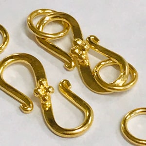May include: Two gold-toned metal S-shaped clasps with a decorative detail on each. The clasps are designed to be used for jewelry making or other craft projects.