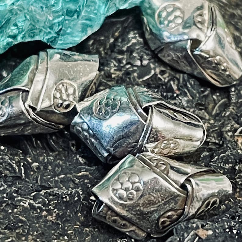 May include: Close-up of a silver metal chain with intricate designs. Each link is shaped like a flower with a raised center. The chain is lying on a dark, textured surface.