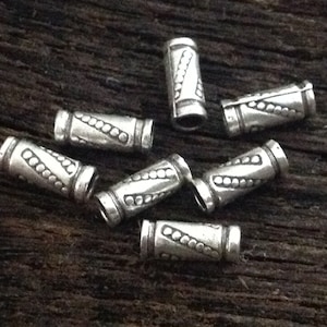 May include: Close-up of several small, silver-colored metal beads. Each bead is cylindrical with a repeating pattern of dots and lines. The beads are scattered on a dark, textured wooden surface.
