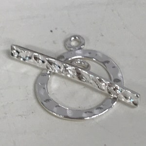 Könnte beinhalten: Silberner Toggle-Verschluss mit einer strukturierten Stange und einem gehämmerten Kreis. Der Verschluss ist für Halsketten, Armbänder oder anderen Schmuck gedacht.