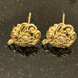 May include: A pair of gold-colored stud earrings. Each earring features an ornate, floral design with intricate details. The earrings are displayed on a reflective surface, creating a mirrored effect. The posts are visible, indicating they are designed for pierced ears.