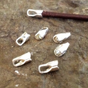 May include: Six silver-toned metal cord ends with a loop on one end and a flat, open end on the other. The cord ends are arranged on a rough, gray surface.
