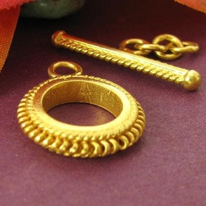 May include: Gold-toned metal toggle clasp with a rope-like design and a round, decorative ring. The ring has a textured, braided edge.