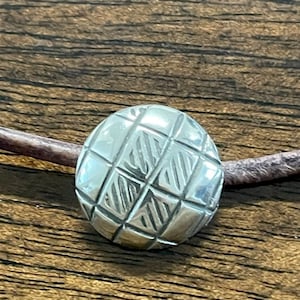 May include: A silver round pendant with a geometric pattern, attached to a brown leather cord.