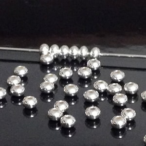 May include: A collection of silver-toned metal beads, some arranged in a line and others scattered on a black surface. The beads are round and have a smooth, reflective finish.
