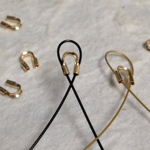 May include: Two gold-toned crimp beads are attached to black and gold cords. The cords are crossed over each other. The crimp beads are used to secure the ends of cords or cables.