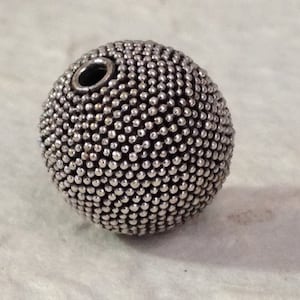 Könnte beinhalten: Eine silberne Perle mit einer strukturierten Oberfläche, hergestellt aus kleinen, runden, metallischen Perlen. Die Perle hat ein Loch in der Mitte zum Aufziehen.