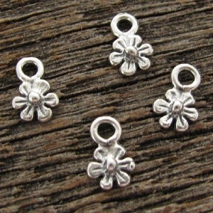 May include: Four small, silver-colored daisy charms with a loop at the top for jewelry making. The charms have a detailed flower design with petals and a central sphere. The charms are displayed on a textured, brown wooden surface.