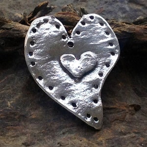 Puede incluir: Un amuleto de plata en forma de corazón con un corazón más pequeño en relieve en el centro. El amuleto tiene una superficie texturizada y un borde punteado.
