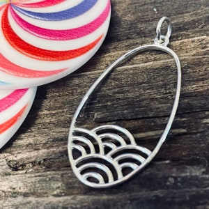 May include: A silver oval pendant with a cut-out design of overlapping waves. The pendant has a loop at the top for attaching a chain.