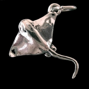 May include: A silver-colored stingray pendant with a curved tail and a loop for attaching to a chain. The pendant is highly detailed and has a polished finish, set against a black background.