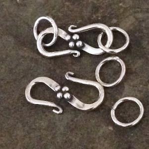 May include: Two silver-toned metal clasps with decorative accents. Each clasp features a curved hook and a small circle. The clasps are designed to be used for jewelry making.