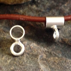 May include: Close-up of silver jewellery findings, including a clasp and a connector. A reddish-brown cord is partially visible, threaded through a silver tube. The findings are on a textured, dark surface.