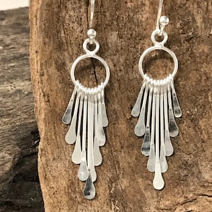 May include: A pair of silver dangle earrings. Each earring features a circular top with a series of vertical, elongated teardrop-shaped dangles. The earrings are suspended from a simple hook. The background is a textured, wooden surface.