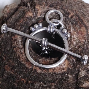May include: A silver toggle clasp with a black circular center. The clasp features a bar with rounded ends and a ring for attachment. The design includes decorative beaded accents around the circular portion.