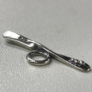 May include: Silver-toned metal charm with a curved shape and a small ring. The charm has a textured surface and three small dots on the end.