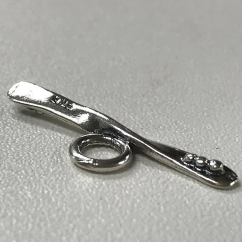 May include: Silver-toned metal charm with a curved shape and a small ring. The charm has a textured surface and three small dots on the end.