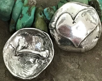 May include: Two silver-toned, irregularly shaped beads. One bead features a heart design, while the other has a curved, bone-like shape. The beads are set against a backdrop of turquoise-colored stones, suggesting a jewelry component or decorative item.