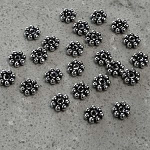 May include: A collection of small, silver-toned metal beads shaped like flowers. Each bead features a central hole surrounded by a ring of tiny, raised spheres, creating a textured, circular design. The beads are scattered across the surface.