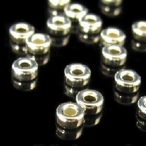 May include: A close-up of a group of silver-toned metal spacer beads. The beads are round with a hole in the center. They are arranged in a random pattern on a black surface.