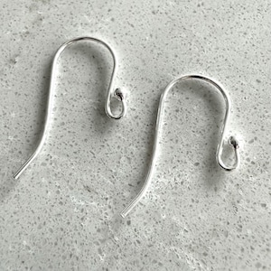 May include: Two silver-colored earring hooks are displayed against a light gray, speckled surface. Each hook has a curved design with a small, rounded end. The hooks are designed for pierced ears and are likely for handmade jewelry.
