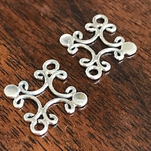 May include: Two silver-colored filigree components with an ornate, symmetrical design. Each component features a central design with curved lines and circular accents. The components are displayed on a dark wooden surface.