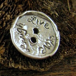 May include: A silver-colored button with the words "Live Your Dream" engraved on its surface. The button has two holes for sewing and a textured, slightly irregular edge. The button is set against a dark, textured background.
