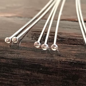 May include: A close-up of a group of silver head pins with clear glass heads. The pins are arranged in a row on a dark brown wooden surface.