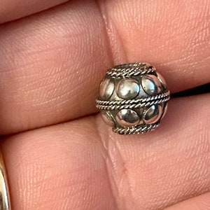 May include: A small, silver bead with a rounded form. It has a pattern of raised, circular designs and is accented with twisted rope-like details. The bead is held in a hand, indicating its size and intricate design.