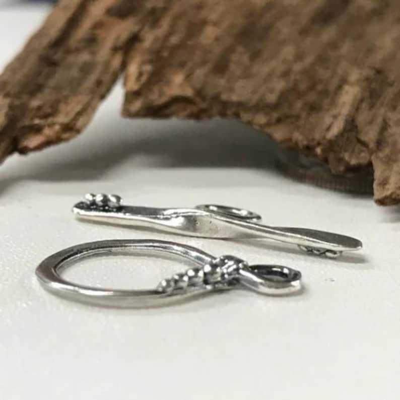 May include: Two silver-toned jewelry findings, one is a simple loop and the other is a flat, elongated shape with a rounded end. Both have a textured design.
