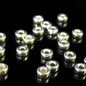 May include: A close-up of a group of silver-toned metal spacer beads. The beads are round with a hole in the center. They are arranged in a random pattern on a black surface.
