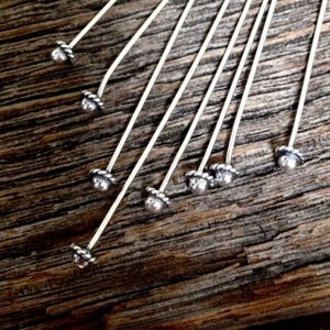 May include: Close-up of several silver-toned head pins with decorative, textured tops. The pins are arranged diagonally against a rustic, weathered wooden surface. The tops have a small, round, silver bead surrounded by a twisted rope design.
