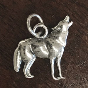 Puede incluir: Un colgante plateado con la forma de un lobo aullando. El lobo está representado de pie con la cabeza levantada y la boca abierta. El colgante tiene un lazo en la parte superior para sujetarlo a una cadena o pulsera. El fondo es una superficie de madera marrón oscuro.